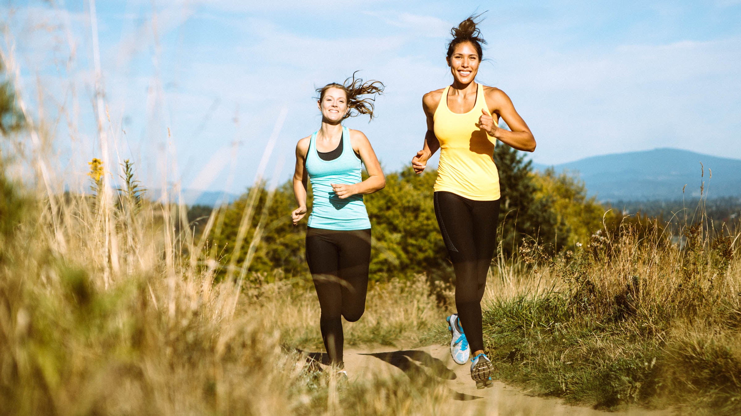 Two women run on a trail in a rural mountain area, smiles on their faces as they enjoy the exercise and warm afternoon.