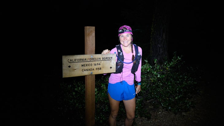 A woman stands next to a California/Oregon border sign at night