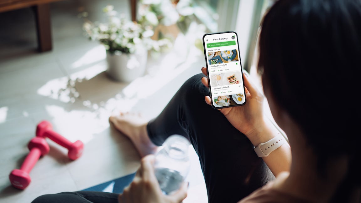 Over the shoulder view of sports woman using meal delivery service and ordering healthy food online with smartphone after exercising at home.