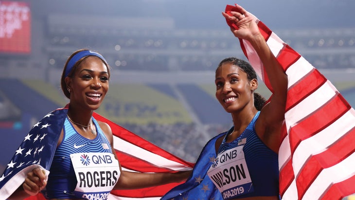 Two women draped with American flags as they wave to fans in a stadium
