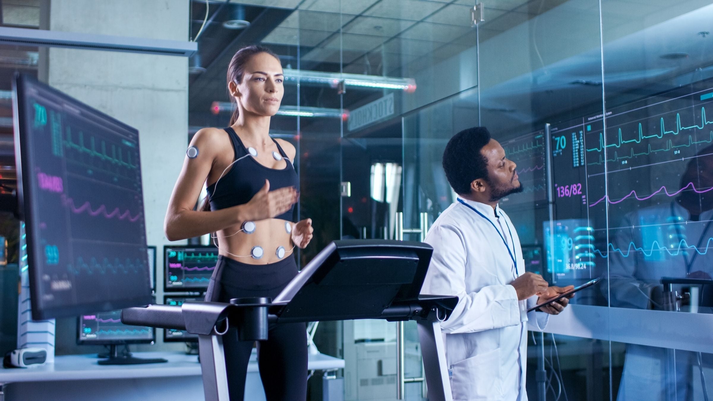 A woman on a treadmill is hooked up to physiology testing equipment. A man in a lab coat observes data.
