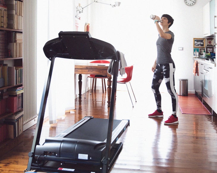A basic black treadmill in the foreground with a woman drinking water in the background