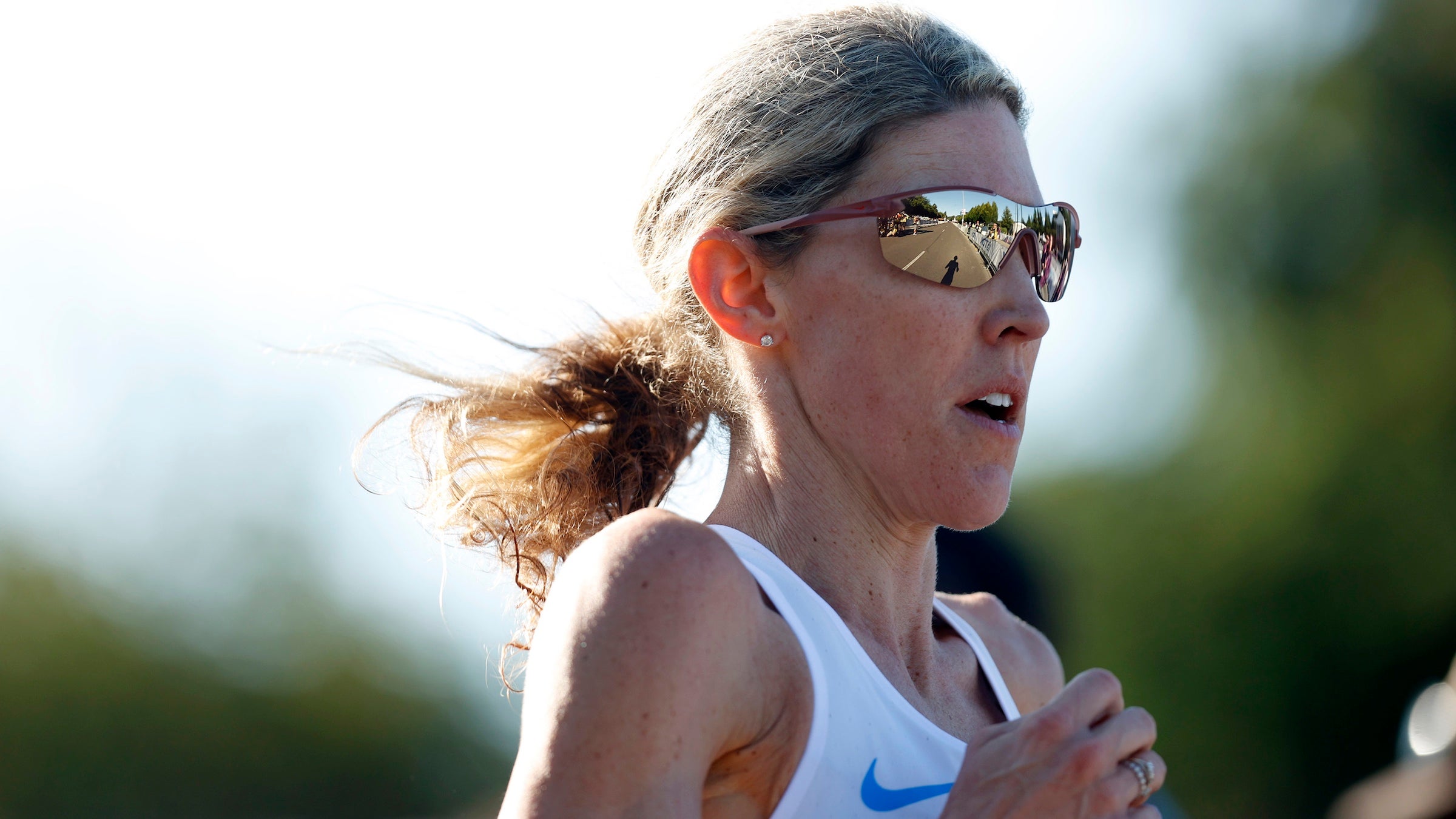 A woman in a white singlet and sunglasses runs a marathon with blonde hair