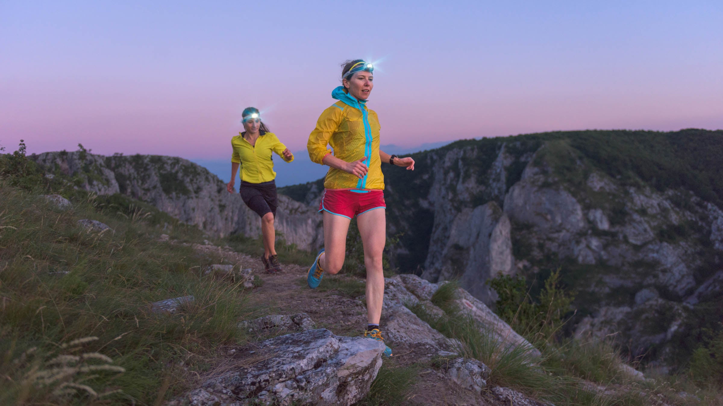 Two female athletes wearing headlamps and running along a hill at sunset